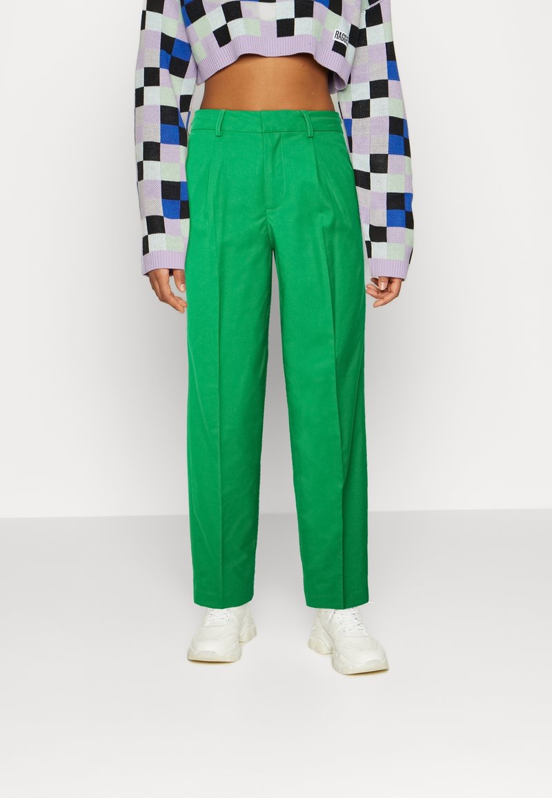 Gröna skräddarsydda byxor med framfållar, gjorda av ett slätt tyg. Stylad med en kort svart och lila rutmönstrad tröja och vita sneakers.