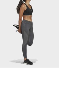Reggiseno sportivo nero e leggings grigi con pannelli testurizzati. Scarpe atletiche nere. Figura che esegue un allungamento con un piede dietro.