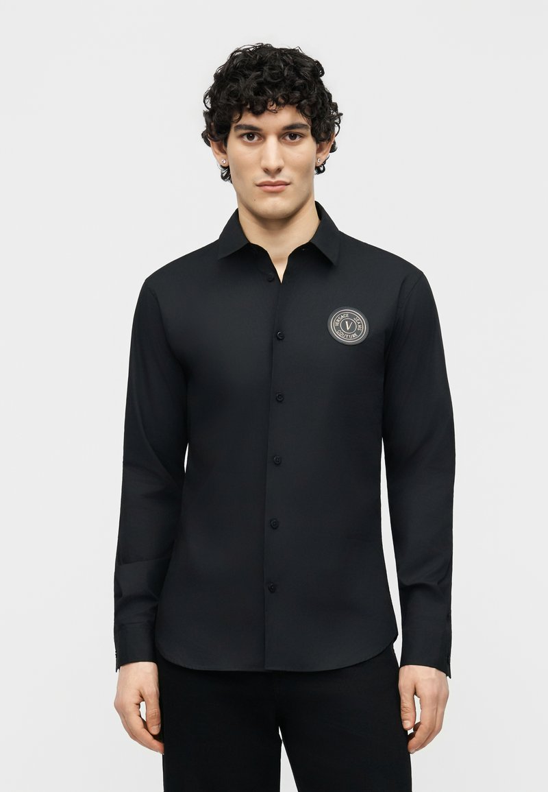 Νέος άνδρας με σγουρά μαλλιά, φορώντας εφαρμοστό μακρυμάνικο μαύρο πουκάμισο με κουμπιά και στρογγυλό έμβλημα στο αριστερό στήθος.