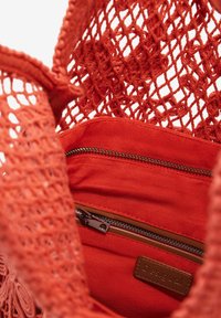 Nærbillede af en rød hæklede taske, der viser en stoflomme med lynlås og et brunt mærkelabel "Desigual".