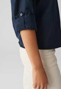 Camicia blu navy con maniche arrotolate realizzata in un tessuto leggero, presenta un polsino con bottone ed è abbinata a pantaloni di colore chiaro.