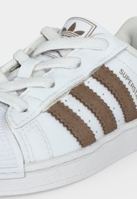 Λευκό αθλητικό παπούτσι Adidas Superstar με καφέ ρίγες, υφασμένα κορδόνια και το λογότυπο Adidas trefoil στη γλώσσα.