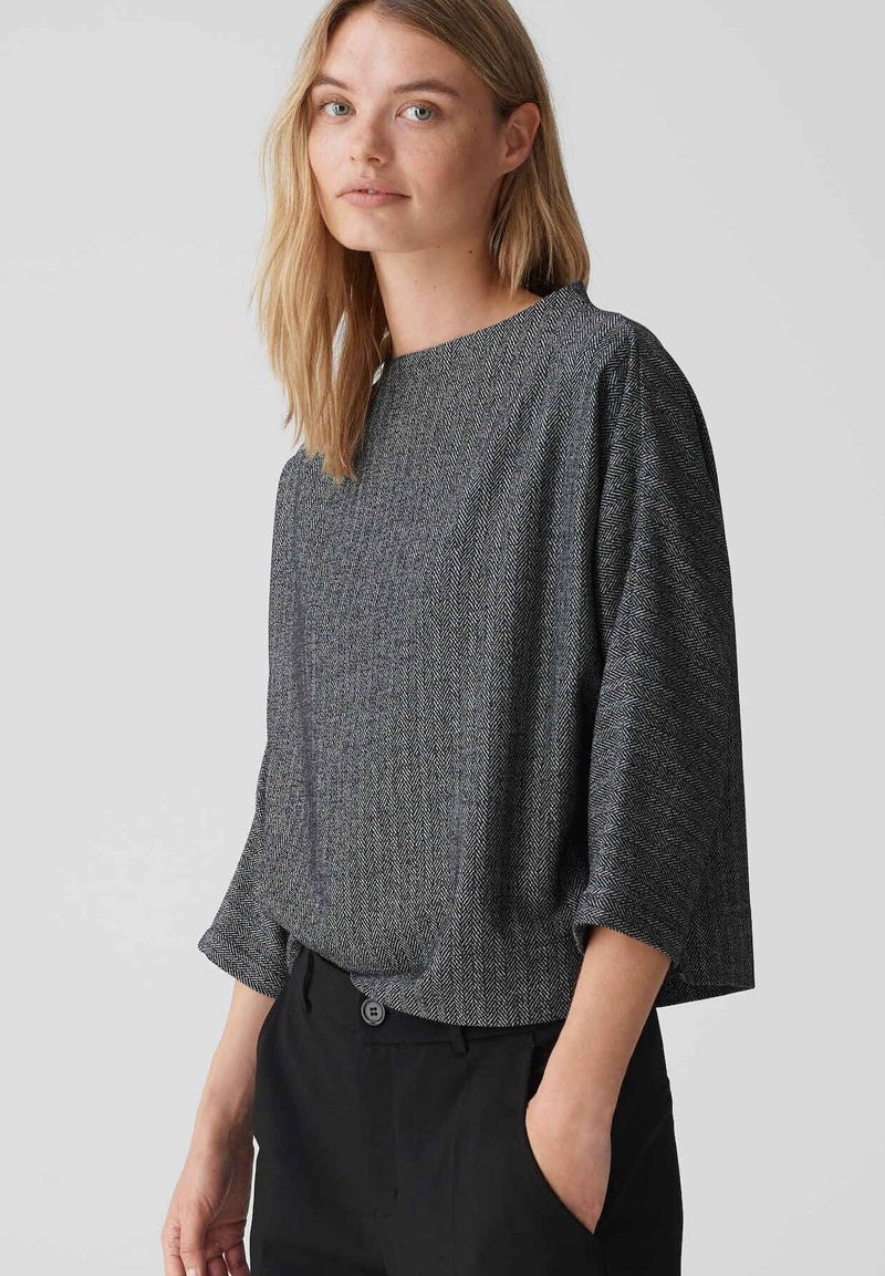 Grijze top met een herringbone-patroon, met een losse pasvorm, ronde hals en driekwartmouwen, gecombineerd met zwarte broek. Zachte textuur.
