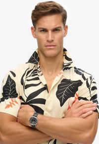 Hombre con cabello corto castaño que lleva una camisa beige con patrones de hojas negras y naranjas, brazos cruzados y un reloj de pulsera de cuero negro.
