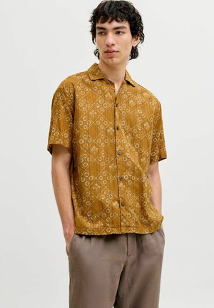Jeune homme aux cheveux bruns bouclés, portant une chemise jaune moutarde à manches courtes avec des motifs floraux et un pantalon marron clair.