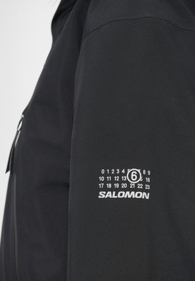 Melna jaka ar gludu tekstūru, ar baltu logotipu "SALOMON" un diagrammu dizainu uz augšējā roka ar skaitļiem no 0 līdz 23.
