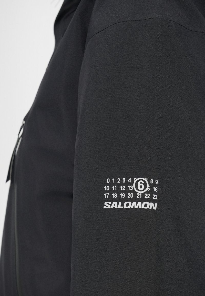 Veste noire à la texture lisse, portant le logo blanc "SALOMON" et un design de graphique sur le bras supérieur avec des numéros allant de 0 à 23.