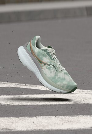 Chaussure de course vert clair et blanc avec des lacets, flottant au-dessus d'une surface en asphalte grise avec des marquages routiers blancs.