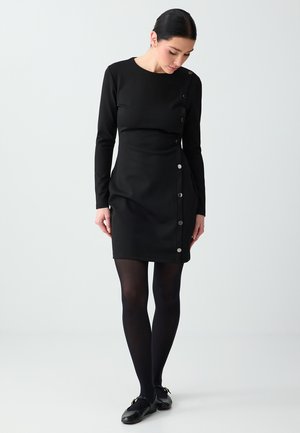 Frau trägt ein schwarzes, langärmliges Kleid mit silbernen Knöpfen an einer Seite, schwarze Strumpfhosen und schwarze Loafers, schaut nach unten.