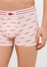 Roze boxershorts van katoenmix, met herhaald patroon van het rode "LAC❤️STE" logo, voorzien van een witte tailleband en rode strepen.