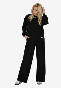 Czarna, oversize bluza z ściągaczami przy mankietach i w pasie, połączona z szerokimi czarnymi spodniami. Model nosi białe trampki i złoty naszyjnik.