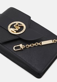 Černá syntetická kabelka s texturovaným povrchem, která je vybavena zlatým kruhovým uzávěrem s logem a řetízkem pro bezpečné zavření.