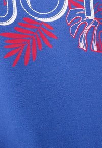 Tela azul con un diseño de costura blanca y patrones de hojas rojas, que muestra una textura suave y un contraste de colores vibrante.