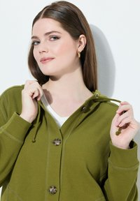 Zelená mikina s kapucí vyrobená z texturované tkaniny s propojením na knoflíky, stahovací kapucí a dvěma vyčnívajícími knoflíky. Obsahuje žebrované manžety a lem.