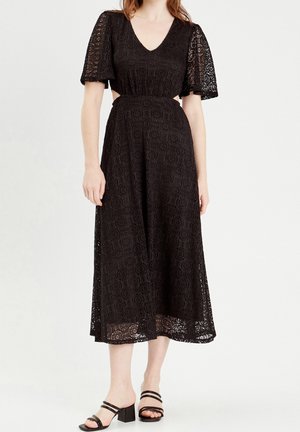 Femme portant une robe midi en dentelle noire avec des manches courtes volantées, un décolleté en V, des côtés découpés et des sandales noires à talons blocs.