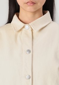 Chemise en velours côtelé beige avec col et boutons en métal argenté, présentant un design texturé en côtes sur le tissu.