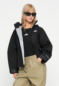 Črna vodoodporna jakna s sivo podlogo, z nastavljivimi manšetami in blagovnimi znamkami. V kombinaciji s črno majico z logotipom in bež hlačami.