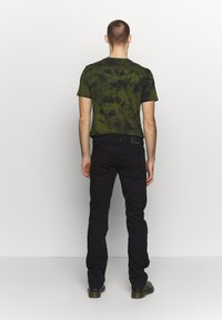 Muž oblečený v zeleno-čiernom tričku s efektom batiky, čiernych džínsoch a čiernych topánkach, stojí čelom k jednoduchým bielym stenám.