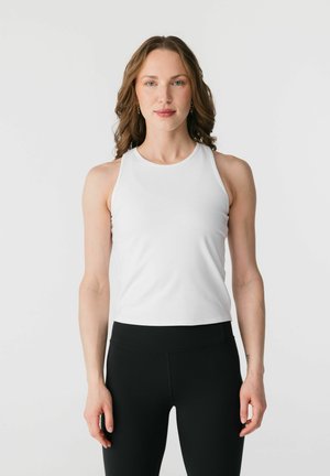 Kvinde med bølget brunt hår iført en ærmeløs hvid top og sorte leggings med høj talje, stående foran en ensfarvet lys baggrund.