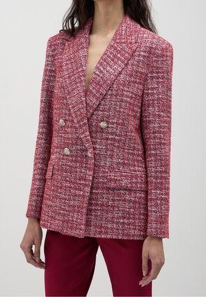 Femme portant un blazer texturé rouge et blanc à double boutonnage avec des boutons argentés, associé à un pantalon rouge uni sur un fond uni.