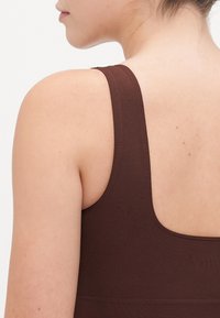 Brown athletic tank top with a high back design, narrow straps, and smooth texture, featuring a subtle pattern. Skin tone visible on shoulder.