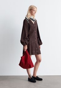 Braunes, strukturiertes Kleid mit langen Ärmeln und Knöpfen, kombiniert mit einer roten Wildledertasche und schwarzen Mokassins. Der Schal hat ein florales Muster.