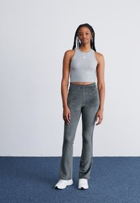 Joven mujer con trenzas largas vistiendo una camiseta sin mangas gris corta, pantalones de tiro alto grises y zapatillas blancas, de pie sobre una alfombra azul.