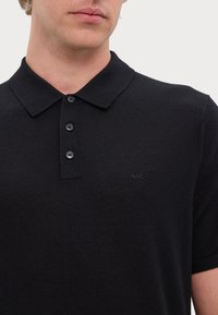 Fekete kötött polo ing, gallérral, három gombos záródással és egy diszkrét logóval hímzett a mellrészen. Rövid ujjak.