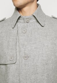 Homme portant un manteau en laine gris clair avec un grand col et quatre boutons visibles, gros plan sur la poitrine et le cou.