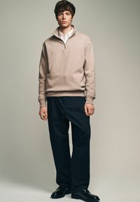 Beige Zip-Up-Sweatshirt mit hohem Kragen, kombiniert mit lockeren, dunkelblauen Hosen und schwarzen Dressschuhen. Glatte Textur, lässige Passform.