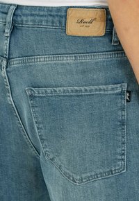 Vaqueros de denim azul claro con un bolsillo trasero, costuras visibles y una etiqueta de cuero marrón claro con la inscripción "Reell" en la cintura.