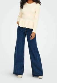 Top peplum crema acanalado con mangas largas combinado con vaqueros azules de pierna ancha y talle alto y zapatos beige. Fondo liso.