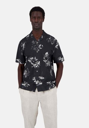 Camicia nera a maniche corte con un motivo floreale bianco, apertura con bottoni e vestibilità rilassata; abbinata a pantaloni in lino beige chiaro.