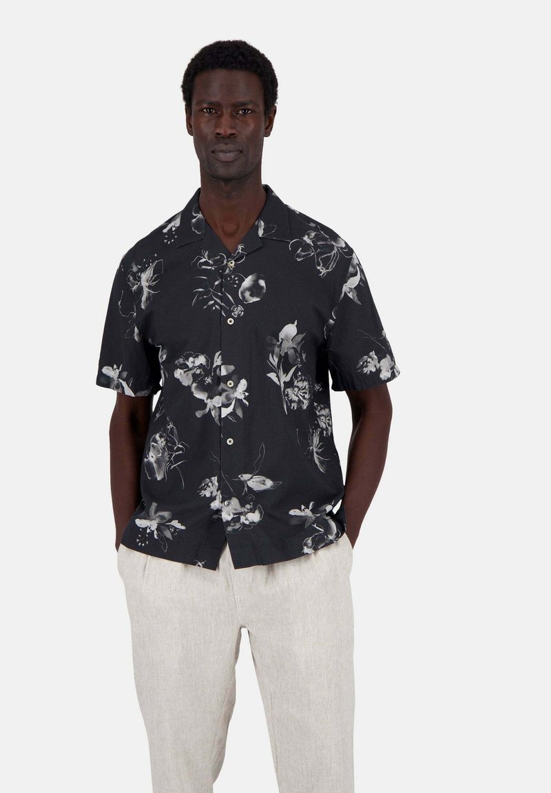 Schwarzes Kurzarmhemd mit einem weißen Blumenmuster, Knopfverschluss und lässigem Schnitt; kombiniert mit hellbeigen Leinenhosen.