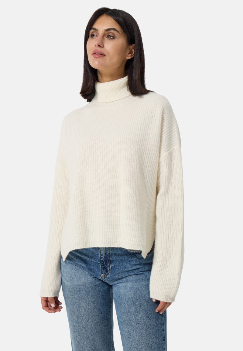 Pull en tricot de couleur crème avec un col haut, texture côtelée et épaules tombantes. Associé à un jean en denim bleu.