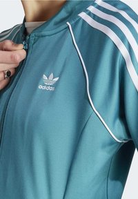 Tyrkysová bunda Adidas se zipem, která má bílé pruhy na rukávech a malou bílou logo na hrudi. Hladká textura.