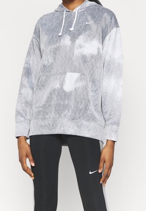 Personne portant un sweat à capuche Nike tie-dye gris et blanc et des leggings Nike noirs avec logos, debout devant un fond clair uni.