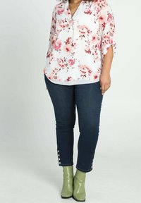 Blusa floreale bianca con accenti rosa, caratterizzata da un dettaglio con cerniera e maniche arrotolabili, abbinata a jeans in denim scuro e stivaletti verdi.