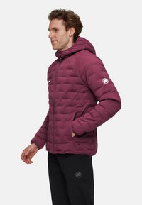 Bordowa kurtka puchowa z kapturem, z zapięciem na zamek z przodu, bocznymi kieszeniami, przeszywanym wzorem i logo na lewej rękawie. Noszona na czarnych spodniach.