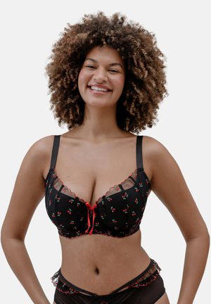 Smiling woman with curly hair wearing black lingerie with red cherry patterns and scalloped lace edges, standing against a plain background.