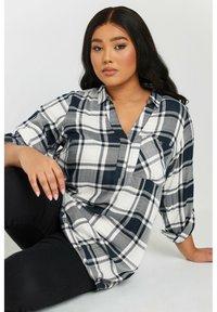 Camisa de cuadros de botones en colores azul marino, gris y blanco; presenta un cuello en V, mangas enrolladas y un bolsillo en el pecho. Textura de tela suave.