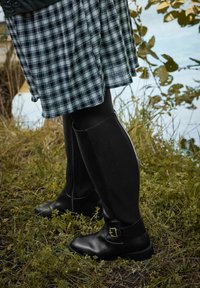 Musta nahkakengät nilkkapituisilla, joissa on solkiyksityiskohta, yhdistettynä ruutuhameeseen ja mustiin sukkahousuihin. Kengissä on sileä pinta ja kestävä muotoilu.