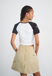 Tricou alb scurt cu mâneci negre și dantelă la margine, asortat cu o fustă din denim bej, cu două buzunare la spate și un patch logo.
