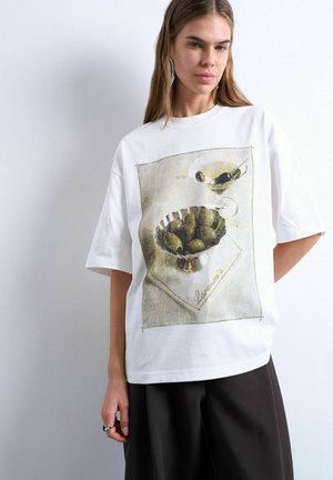 Kvinde iført hvid, oversize T-shirt med trykt billede af oliven i en skål og en martiniglas, stylet med sorte bukser.
