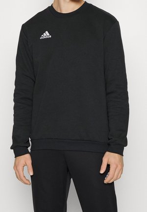 Osoba ubrana w czarną bluzę Adidas oraz dopasowane czarne spodnie, stojąca z rękami swobodnie opuszczonymi wzdłuż ciała.