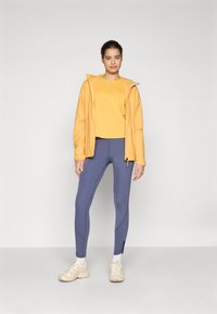 Veste légère jaune sur un haut à manches courtes assorti, associée à des leggings bleus ajustés et des chaussures de sport beiges.