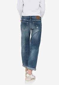 Jeans de mezclilla con un color azul deslavado, detalles desgastados, cinco bolsillos y dobladillos vuelta. La vista trasera muestra una etiqueta de marca de cuero marrón.