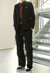 Camicia di velluto a coste nera e pantaloni abbinati con un logo discreto. L'outfit presenta un maglione arancione con motivo sotto e sneakers nere.