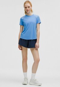 Blå idrottströja med korta ärmar och rund halsringning, kombinerad med marinblå shorts. Modellen bär vita sneakers och randiga strumpor.