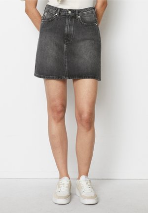 Vrouw die een vervaagde zwarte denim minirok en witte sneakers draagt, staand tegen een witte muur met handen achter de rug.